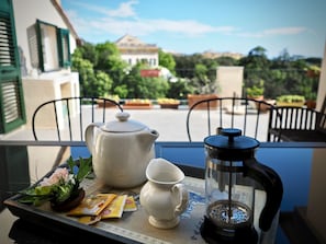 Un vassoio con teiera, caffettiera, tazza e qualche snack su un balcone con vista su un giardino e un edificio.