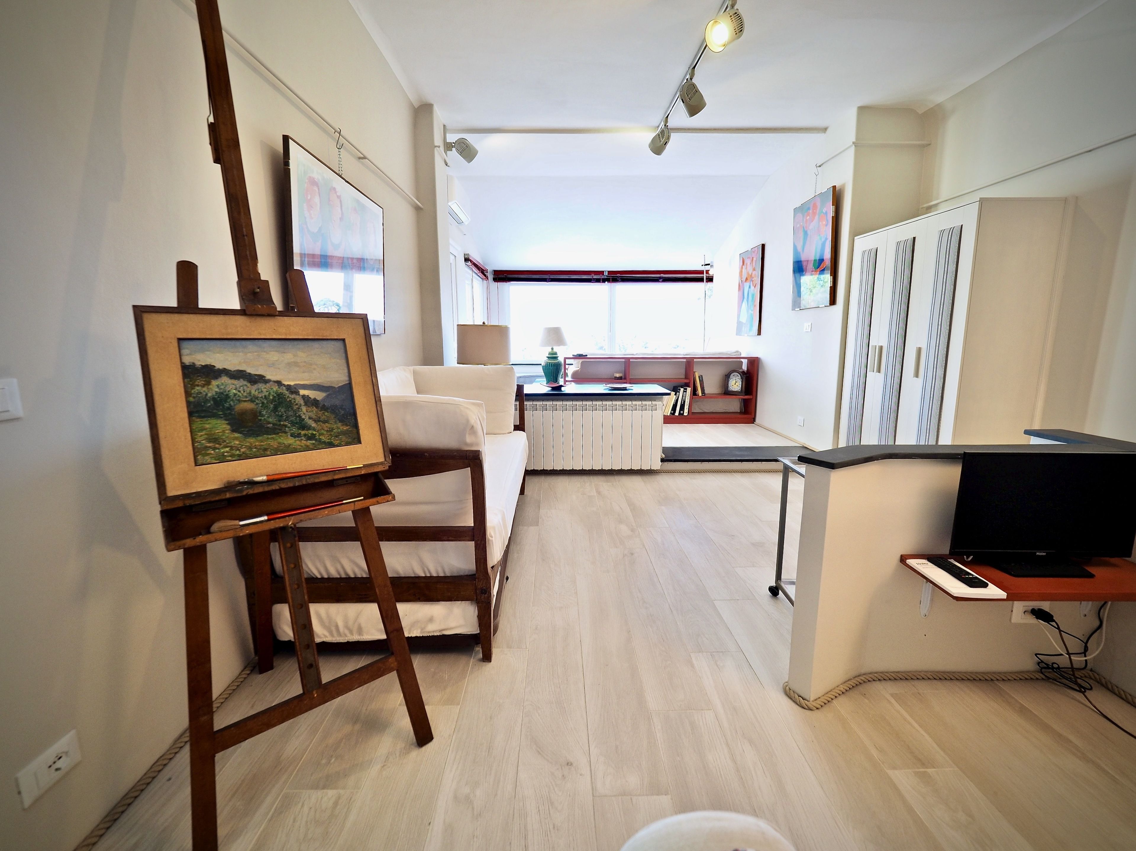 A spacious living area with a wooden easel holding a painting, a white sofa, a small table with a lamp, and a TV stand with a flat-screen TV.