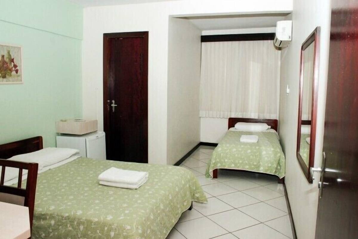 Una habitación de hotel pequeña con dos camas individuales, un escritorio, una silla, un refrigerador pequeño y una puerta que da a otra habitación.