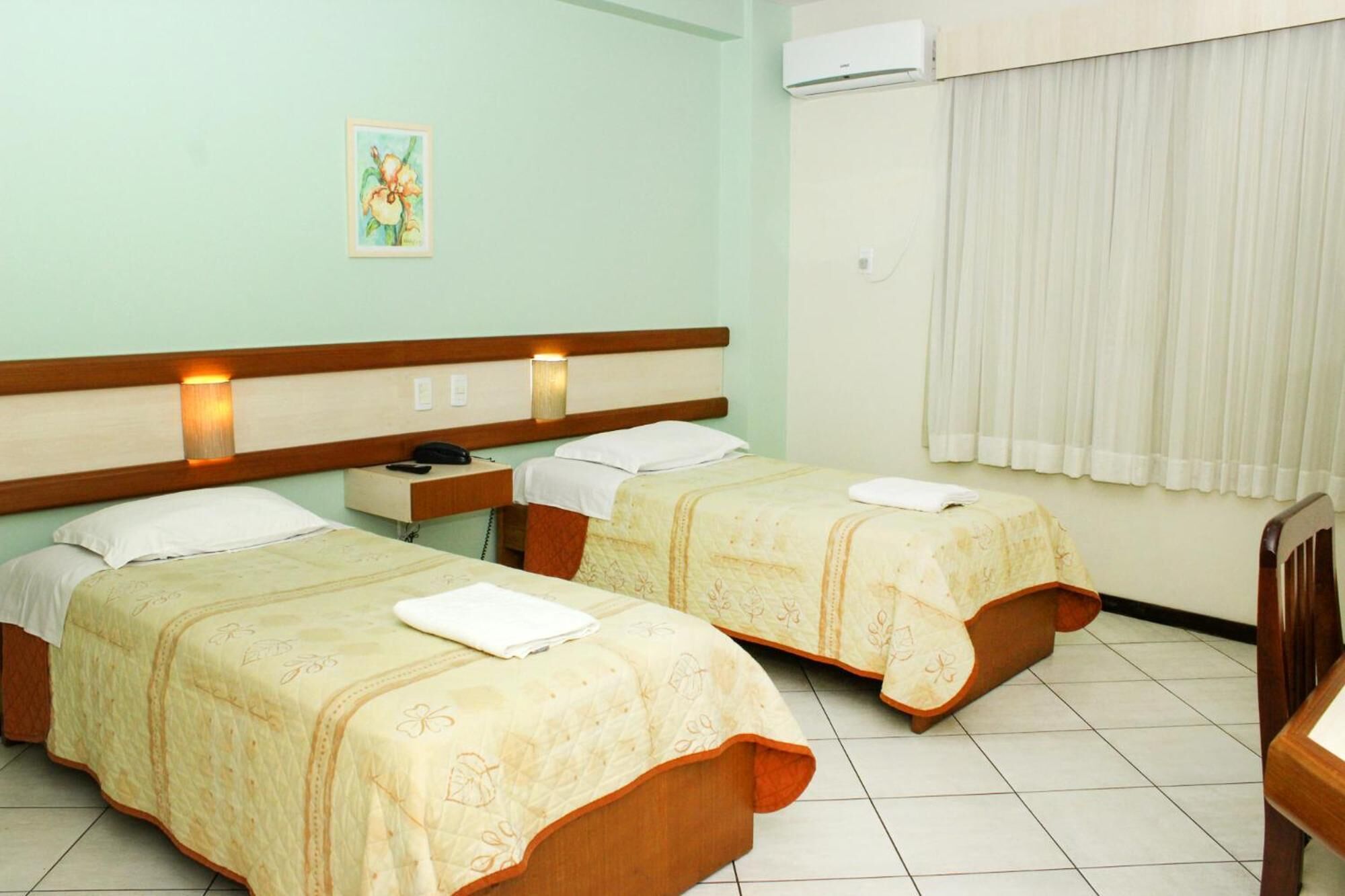 A hotel room with two single beds, a bedside table with a lamp, a painting on the wall, and an air conditioning unit.