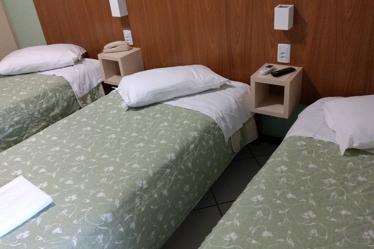 Habitación de hotel con tres camas individuales, cada una con cubrecama verde con estampado floral y almohadas blancas. Hay mesitas de noche con un teléfono y un control remoto.
