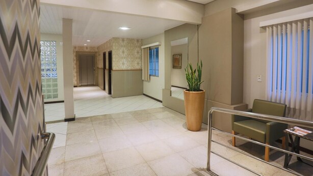 A hotel lobby with a tiled floor, a large potted plant, a seating area with chairs, and a reception desk. - Hotel Veneza (Brusque)