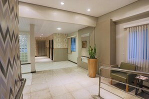 Un hall d’hôtel avec un sol carrelé, une grande plante en pot, un coin salon avec des fauteuils et un comptoir d’accueil.