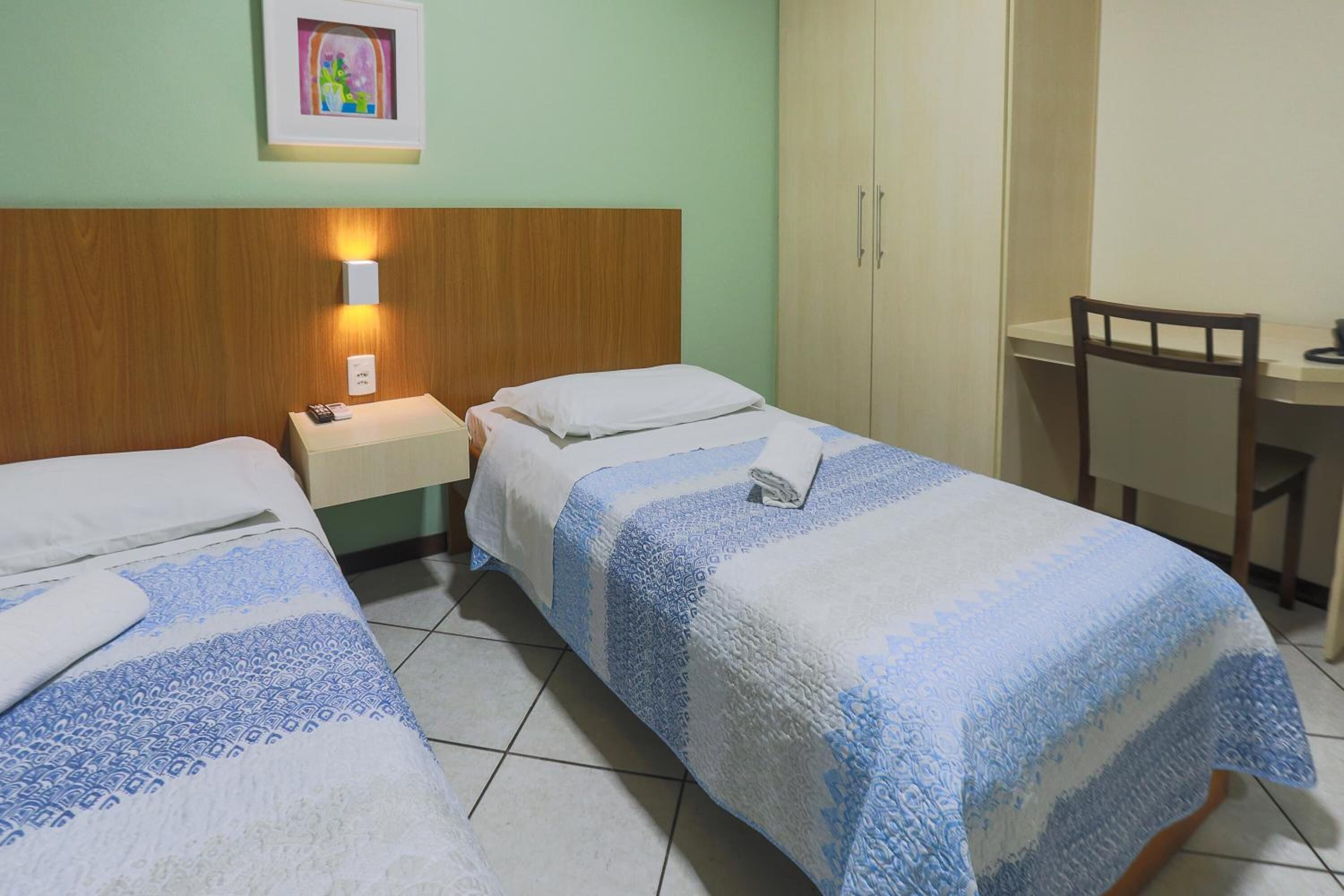 A hotel room with two single beds, a wooden headboard, a bedside table with a lamp, a desk with a chair, and a framed picture on the wall.