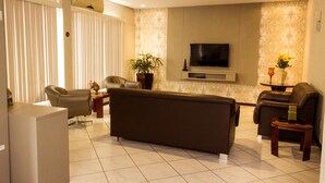 A modern living room with a large sofa, two armchairs, a TV, and a potted plant. - Hotel Veneza (Brusque)