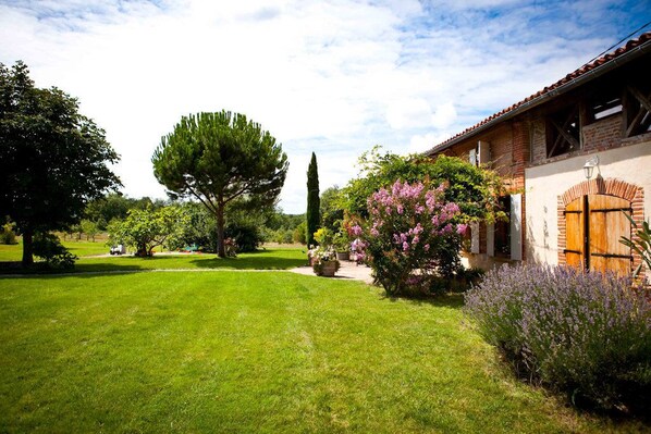 A well-maintained garden with a variety of trees and flowering plants, a clear sky, and a traditional building with a brick facade and wooden door.