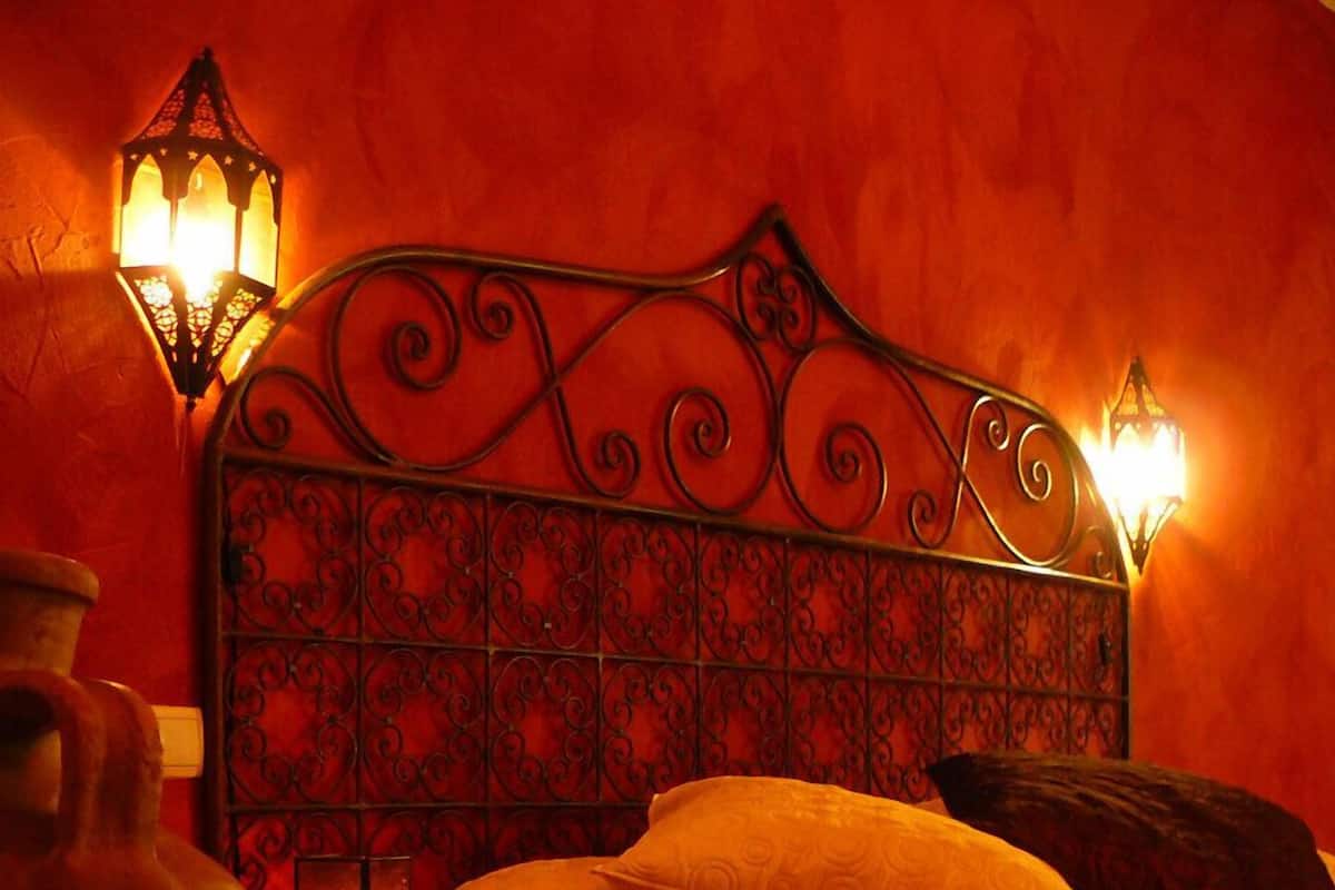 A room with a bed, two wall-mounted lamps, a nightstand with a lamp, a decorative metal headboard, and a patterned rug.