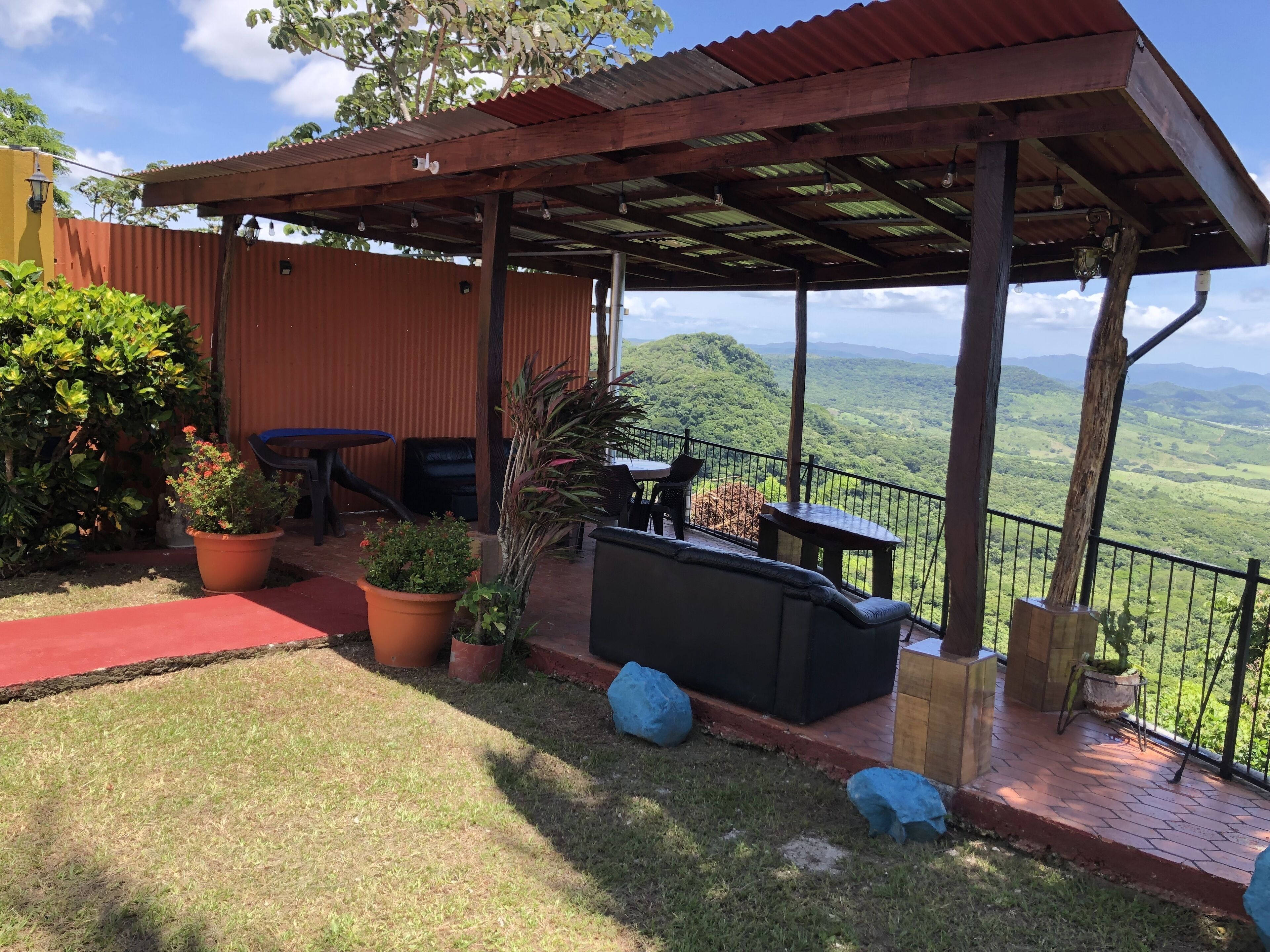 Un patio techado con mobiliario exterior, plantas en macetas y vistas a un valle exuberante.