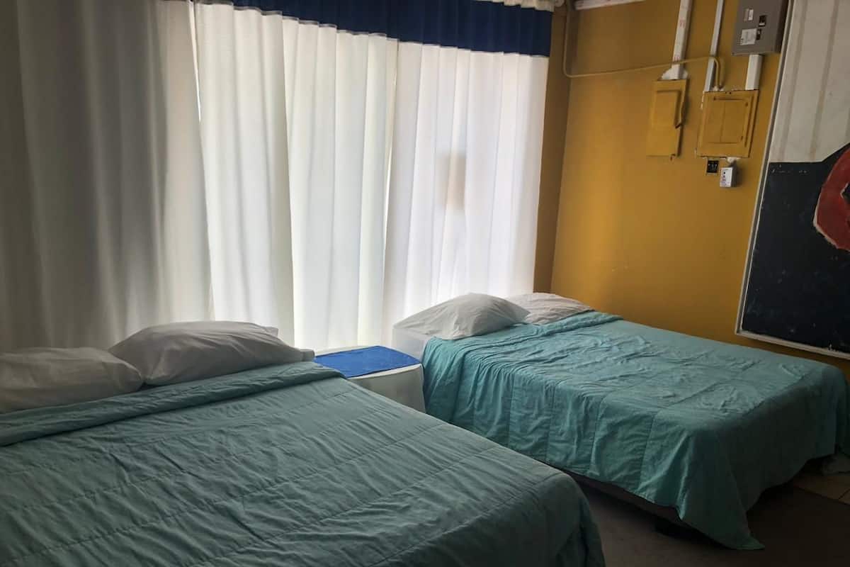 A hotel room with two beds, white curtains, a yellow wall, and a small desk with a blue chair.