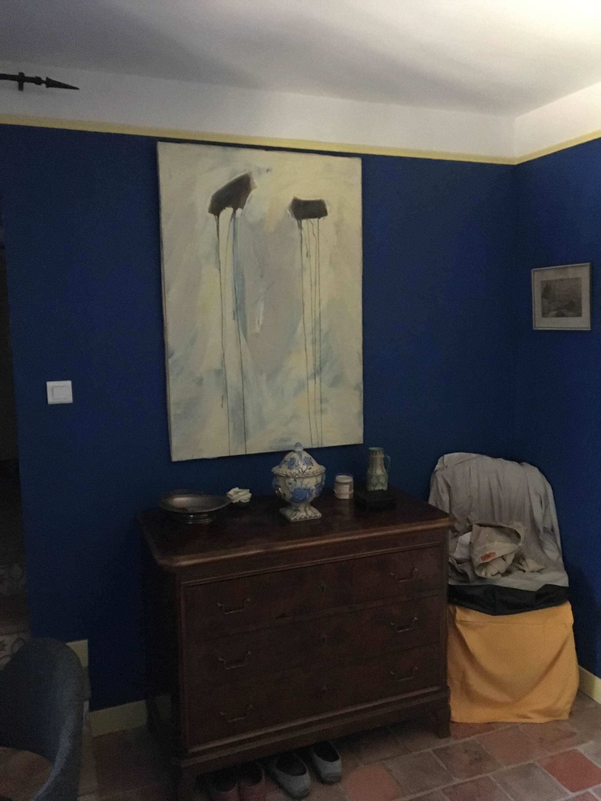 A room with blue walls, a large abstract painting, a wooden dresser with a vase, and a chair.