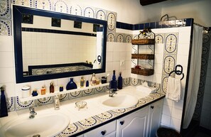 A bathroom with two sinks, a large mirror, and decorative tiles. There are shelves with toiletries and a towel rack. - Domaine du Commandeur (Mondragon)