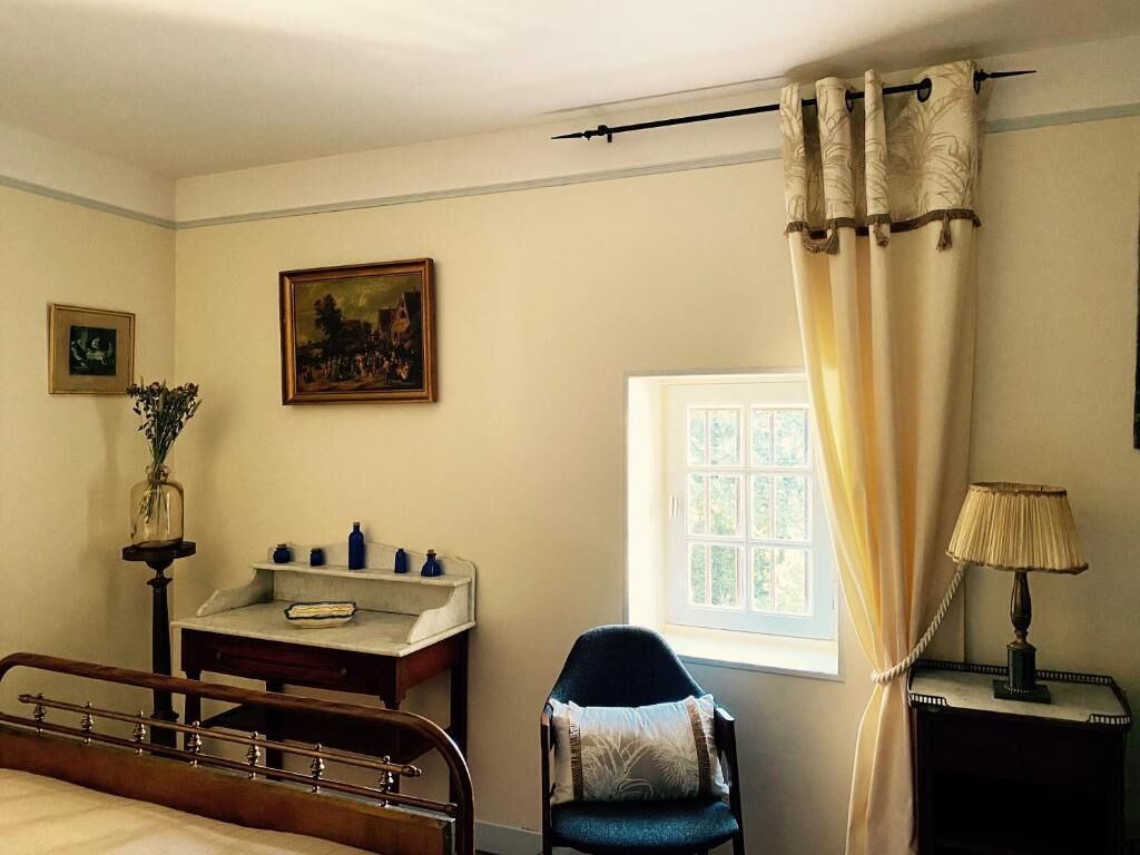 Un dormitorio con cama, una silla, un escritorio con lámpara, una ventana con cortinas y un cuadro en la pared.