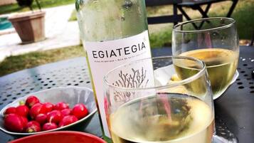 A bottle of Egiategia wine, two glasses of white wine, a bowl of red berries, and a bowl of potato chips on a table outdoors.