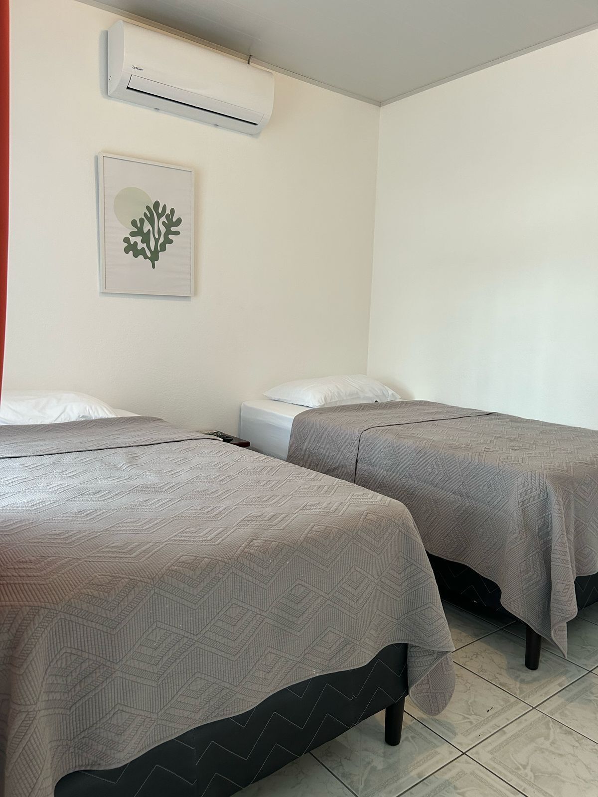 Two single beds with patterned bedding, a wall-mounted air conditioner, and a framed artwork on the wall.