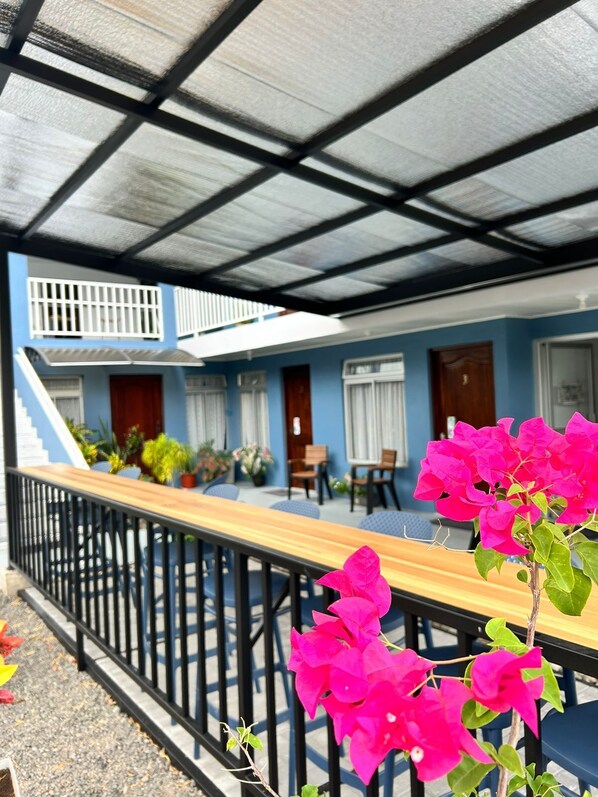 A covered outdoor seating area with a wooden table and chairs, a black metal railing, and a staircase leading to an upper level.
