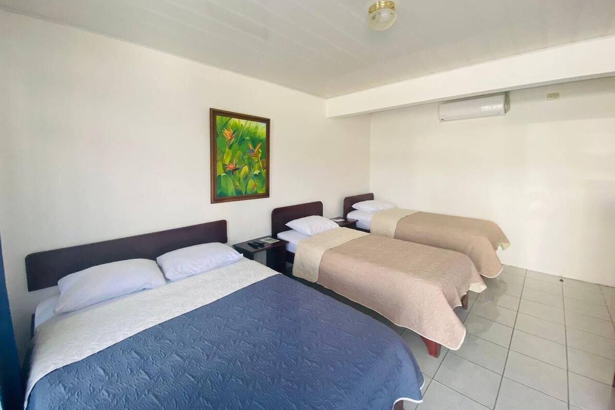 Une chambre d’hôtel avec trois lits simples, une table de chevet avec une lampe, un tableau au mur et un luminaire au plafond.