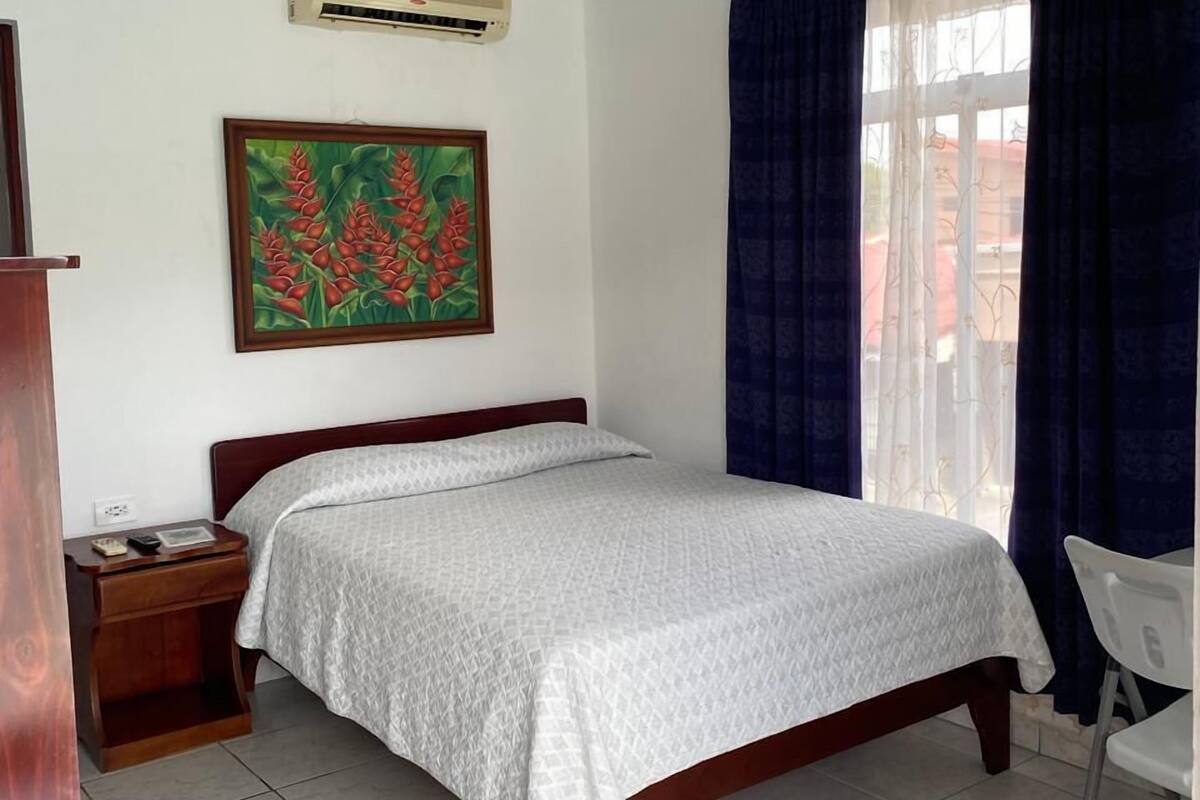 Une chambre à coucher comprenant un lit, une table de chevet, une chaise, un appareil de climatisation et un tableau accroché au mur.