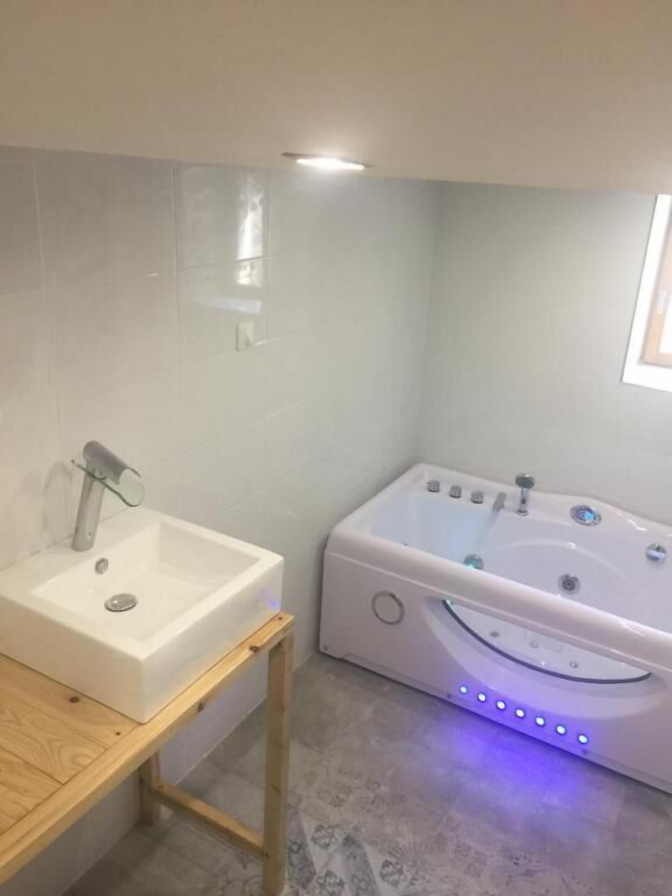Un baño con un lavamanos blanco, una bañera con luces LED y un banco de madera.