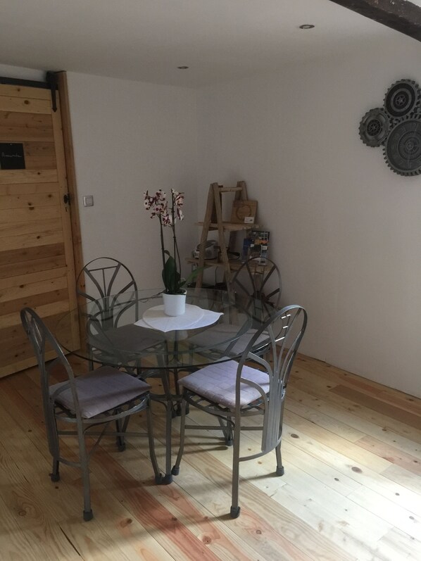 A dining area with a glass table and metal chairs, a wooden floor, and a wall-mounted shelf with books and decorative items.