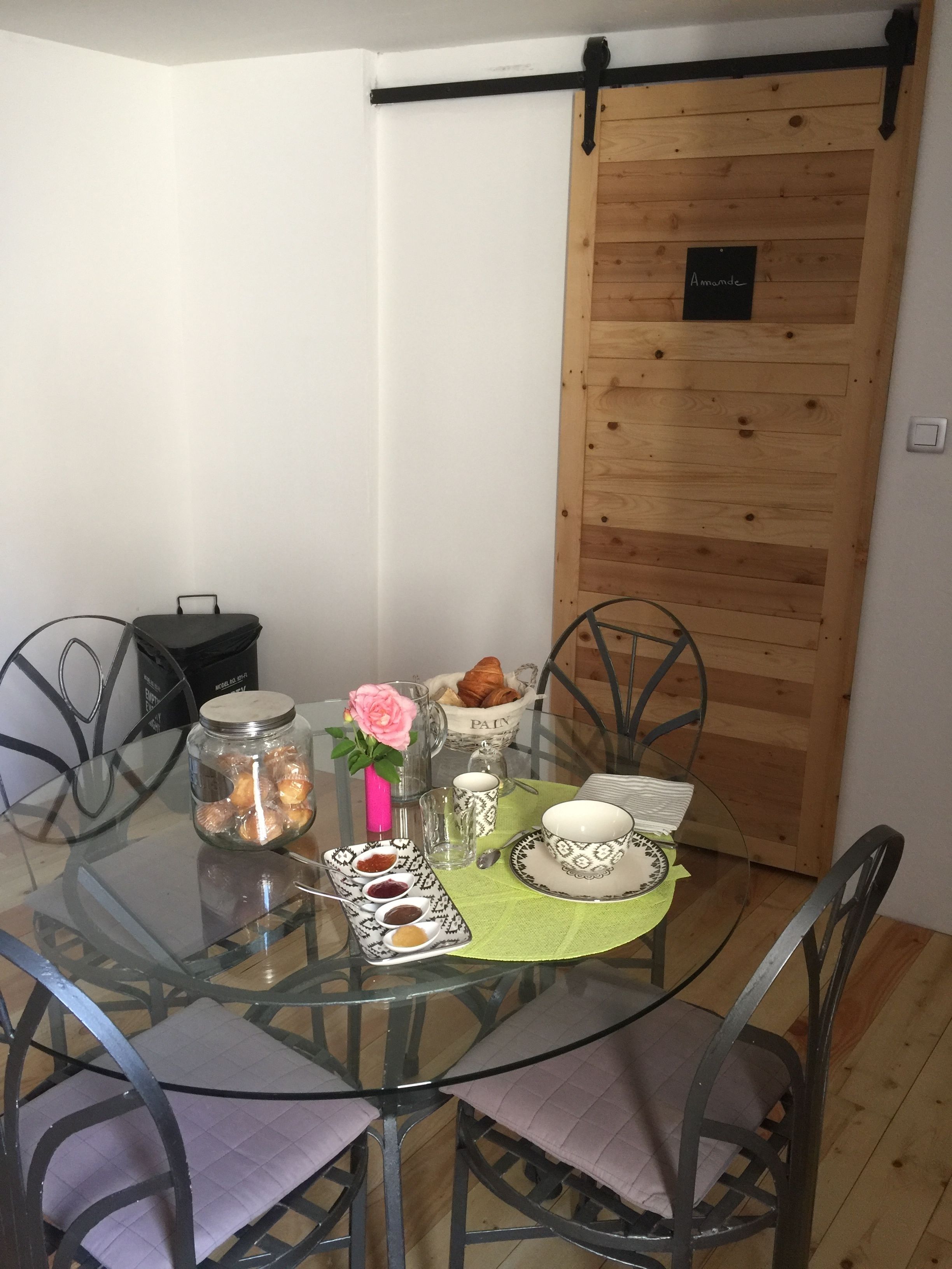Una mesa de comedor con cubierta de vidrio rodeada de sillas de metal, una puerta de madera con un letrero y una jarra de vidrio con una flor rosa.