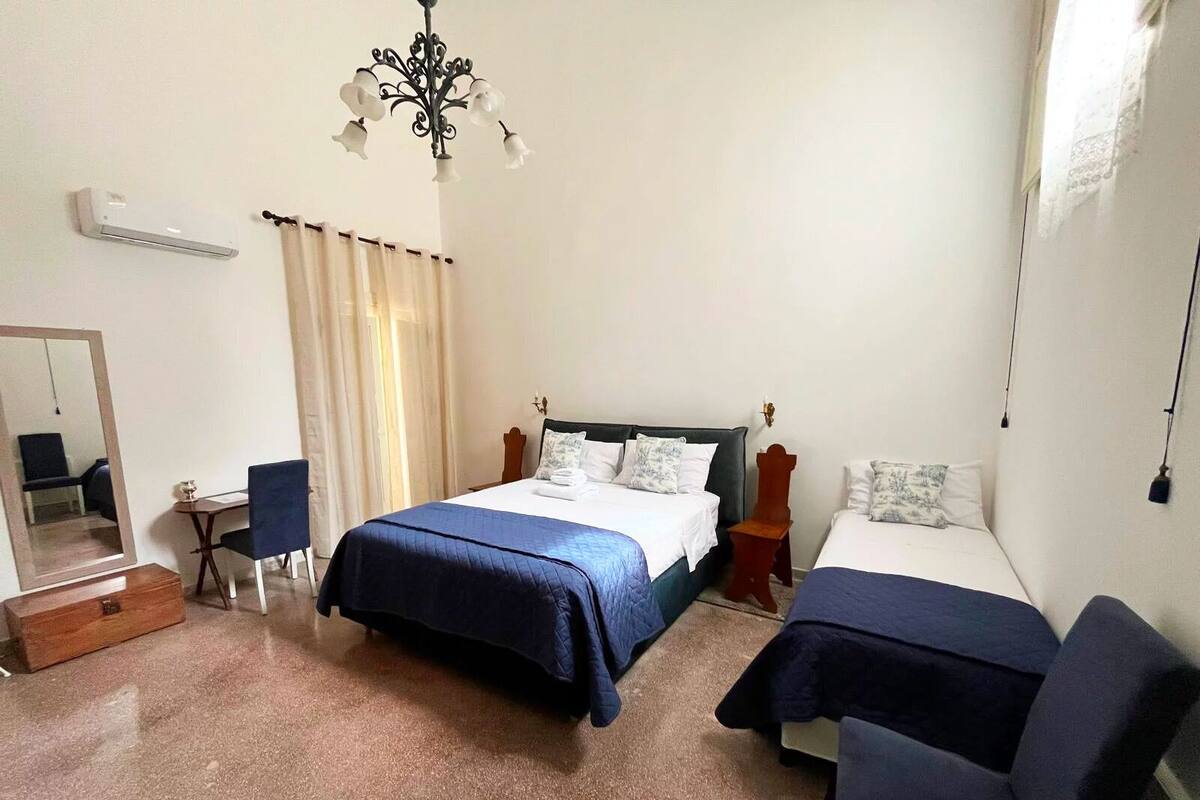 Quarto de hotel com uma cama grande, uma cama menor, uma escrivaninha, uma cadeira, um espelho e um lustre.