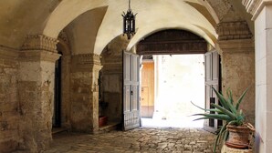 An arched corridor with stone flooring, a potted plant, and an open door leading to a brighter area. - Palazzo Sambiasi (Lecce)