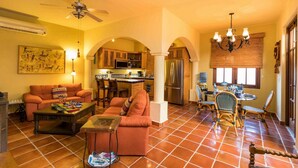 Una sala de estar con sofá, mesa de centro y una zona de comedor con sillas y mesa. Se ve una cocina al fondo, con un refrigerador y un microondas.