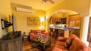Una sala de estar con sofá, mesa de centro y televisor.  Incluye una zona de cocina con microondas y alacenas.