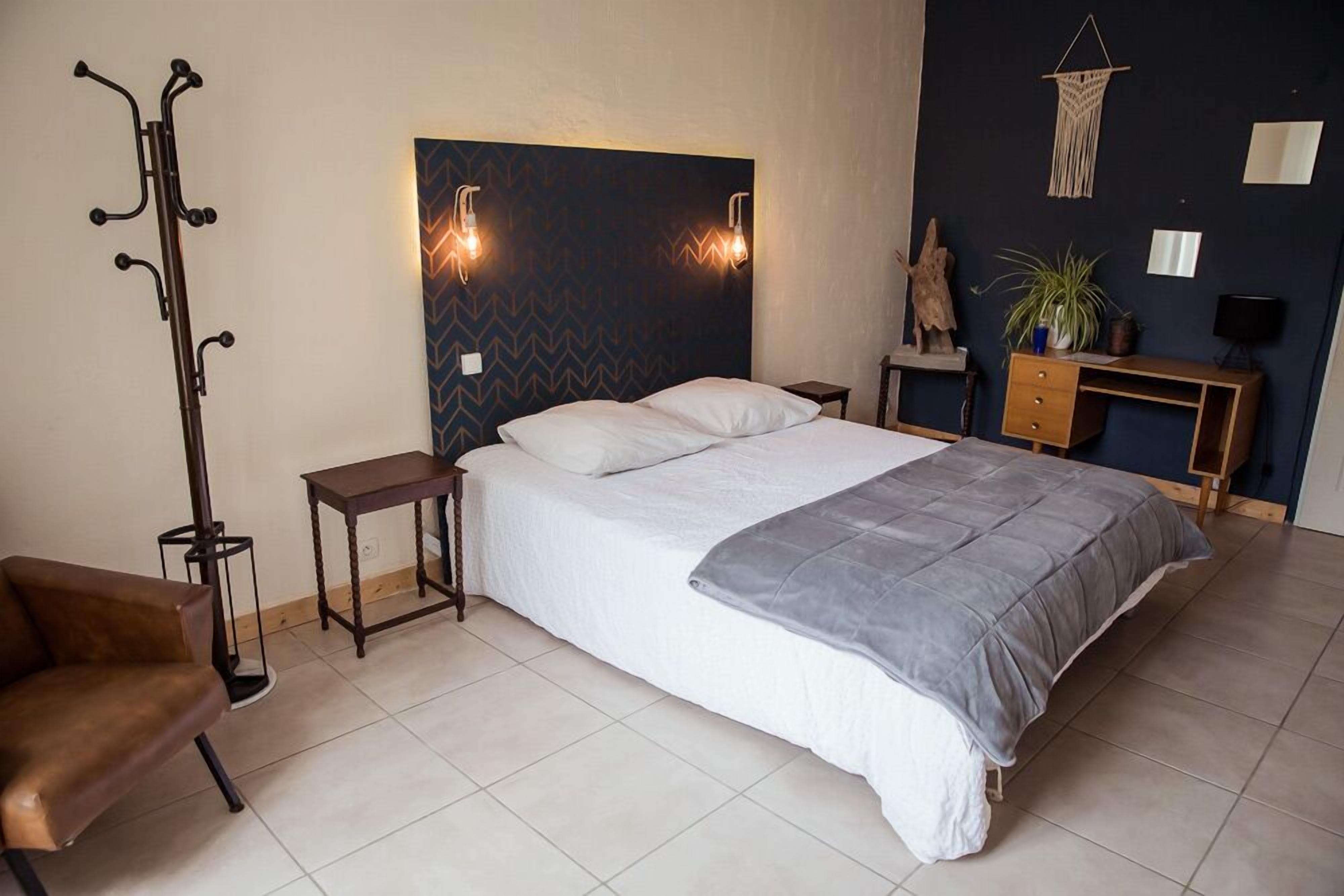 A bedroom with a bed, a coat rack, a desk, a chair, and a plant.