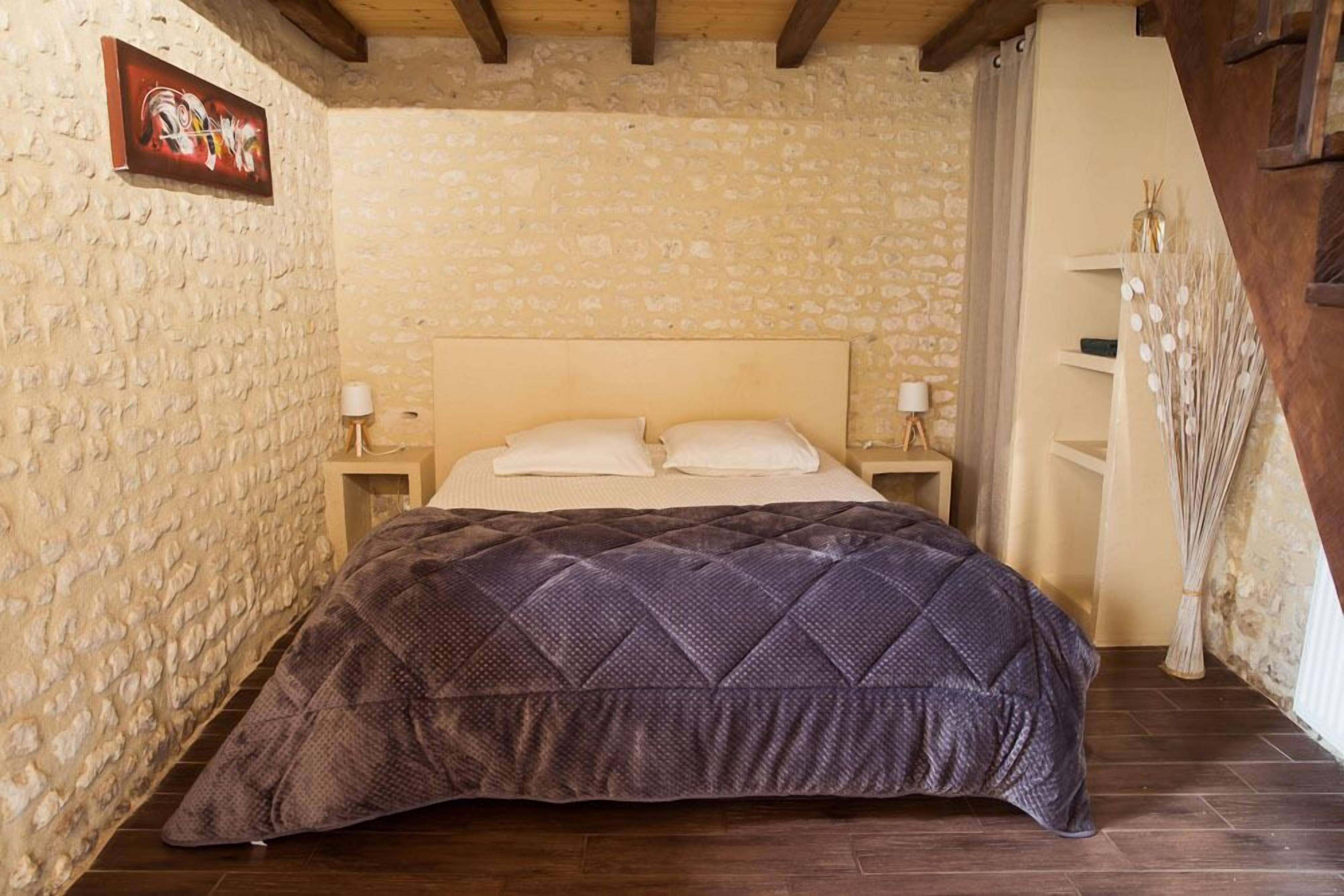 Un dormitori amb paret de pedra, sostre de fusta, un llit amb mantell acolxat, dues taules de nit amb làmpades i una escala de fusta.