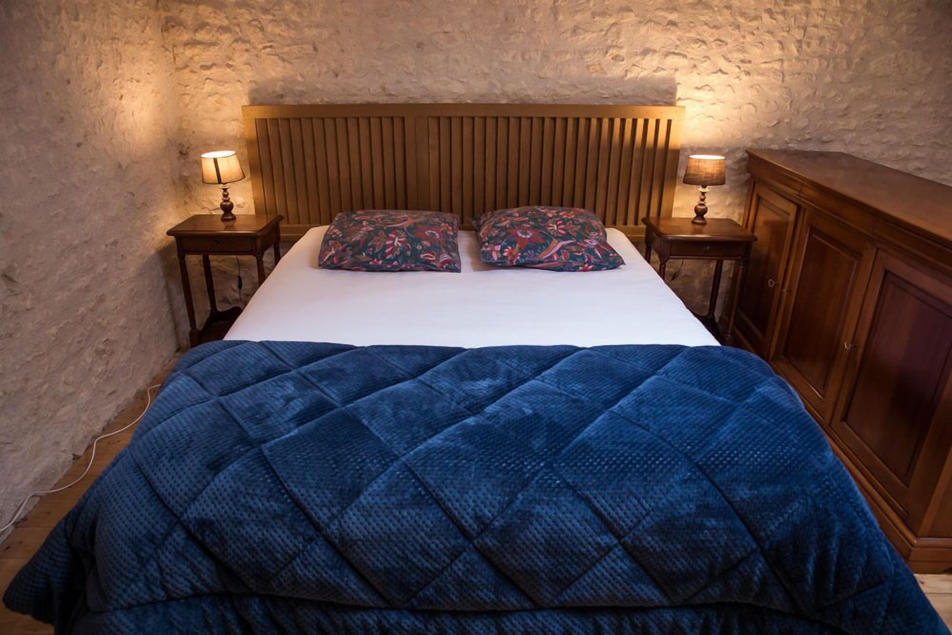 Une chambre à coucher avec une tête de lit en bois, une couvre-lit bleue matelassée, deux coussins à motifs et des lampes de chevet.