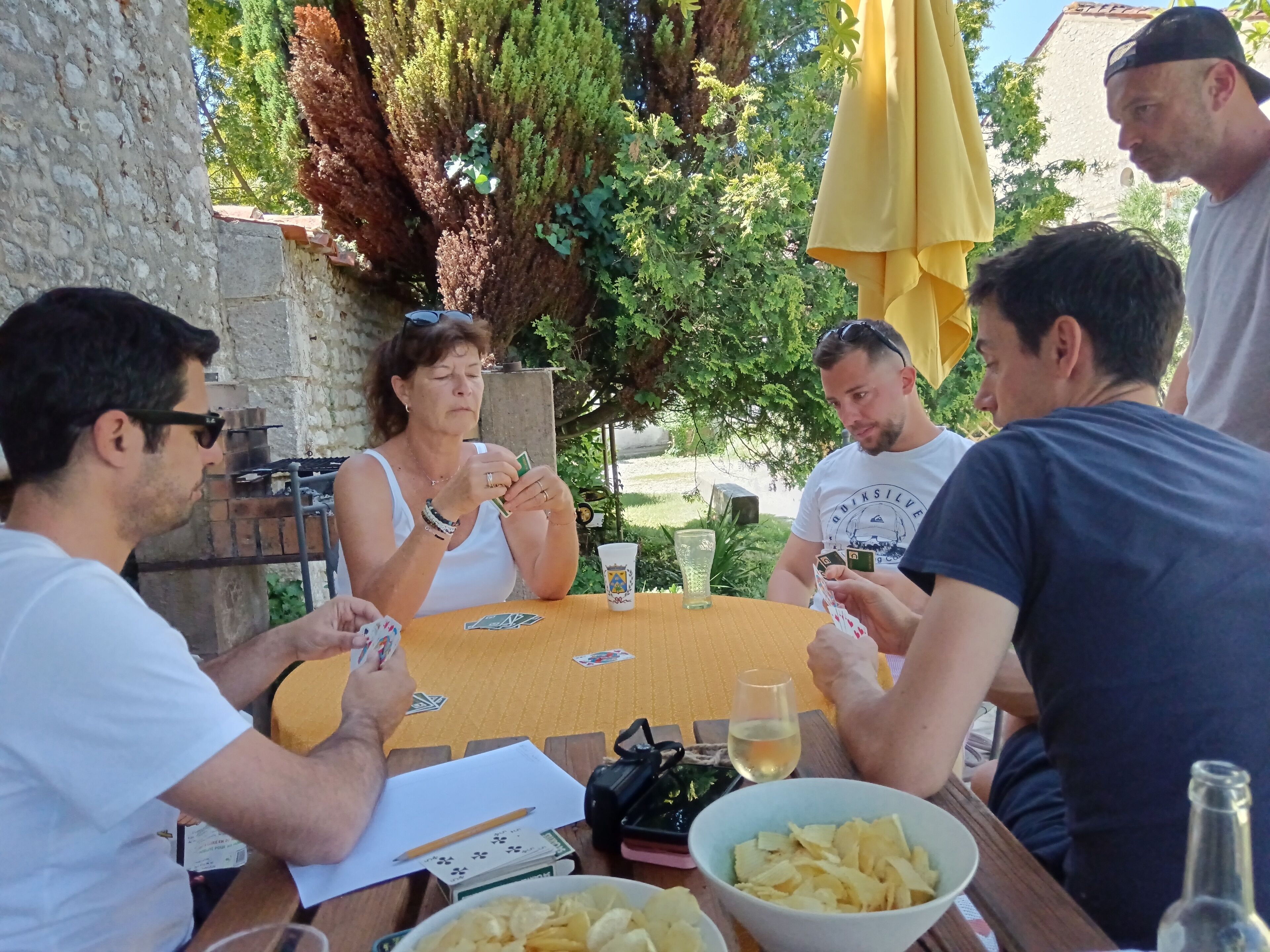 A group of people playing cards at an outdoor table with a yellow umbrella, surrounded by greenery and a stone wall.