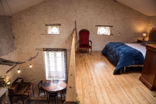 Une pièce rustique avec un plancher en bois, un lit recouvert d’une couverture bleue, une chaise rouge, une table en bois avec des chaises et une petite fenêtre avec un rideau.
