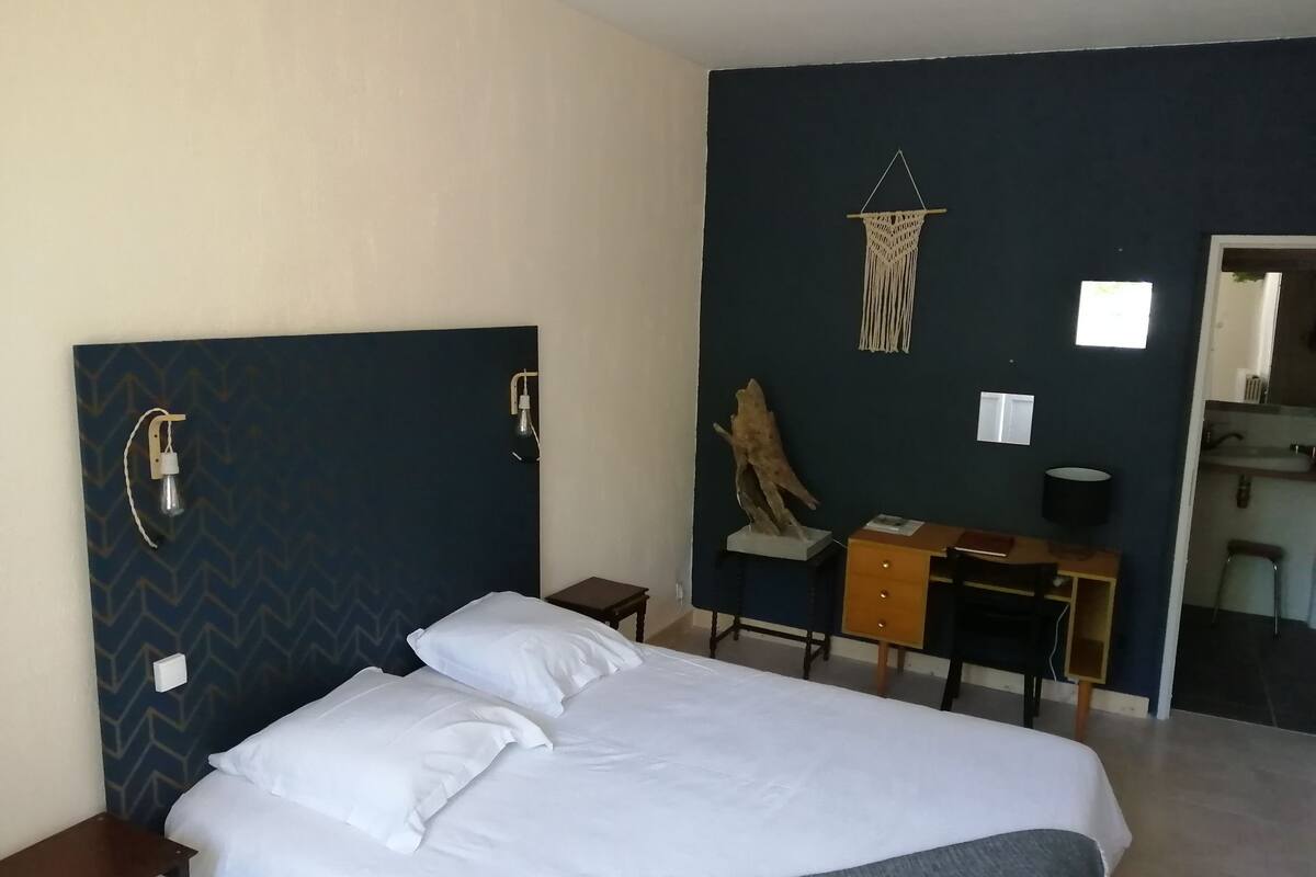 A hotel room with a bed, a desk, a chair, a wall-mounted macrame, and a mirror.