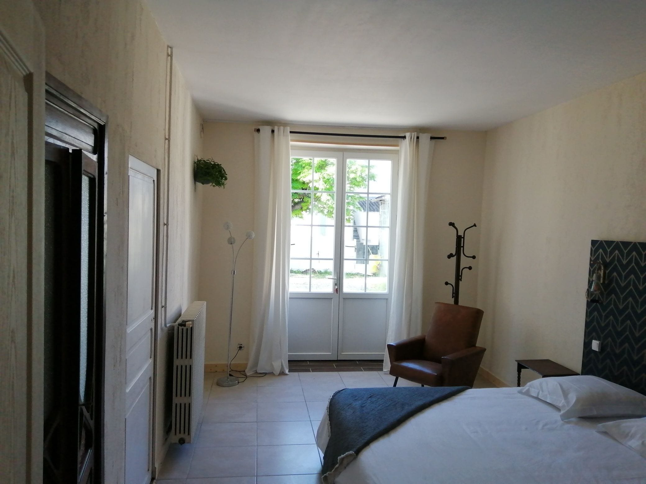 A bedroom with a bed, a chair, a radiator, a window with curtains, and a coat rack.