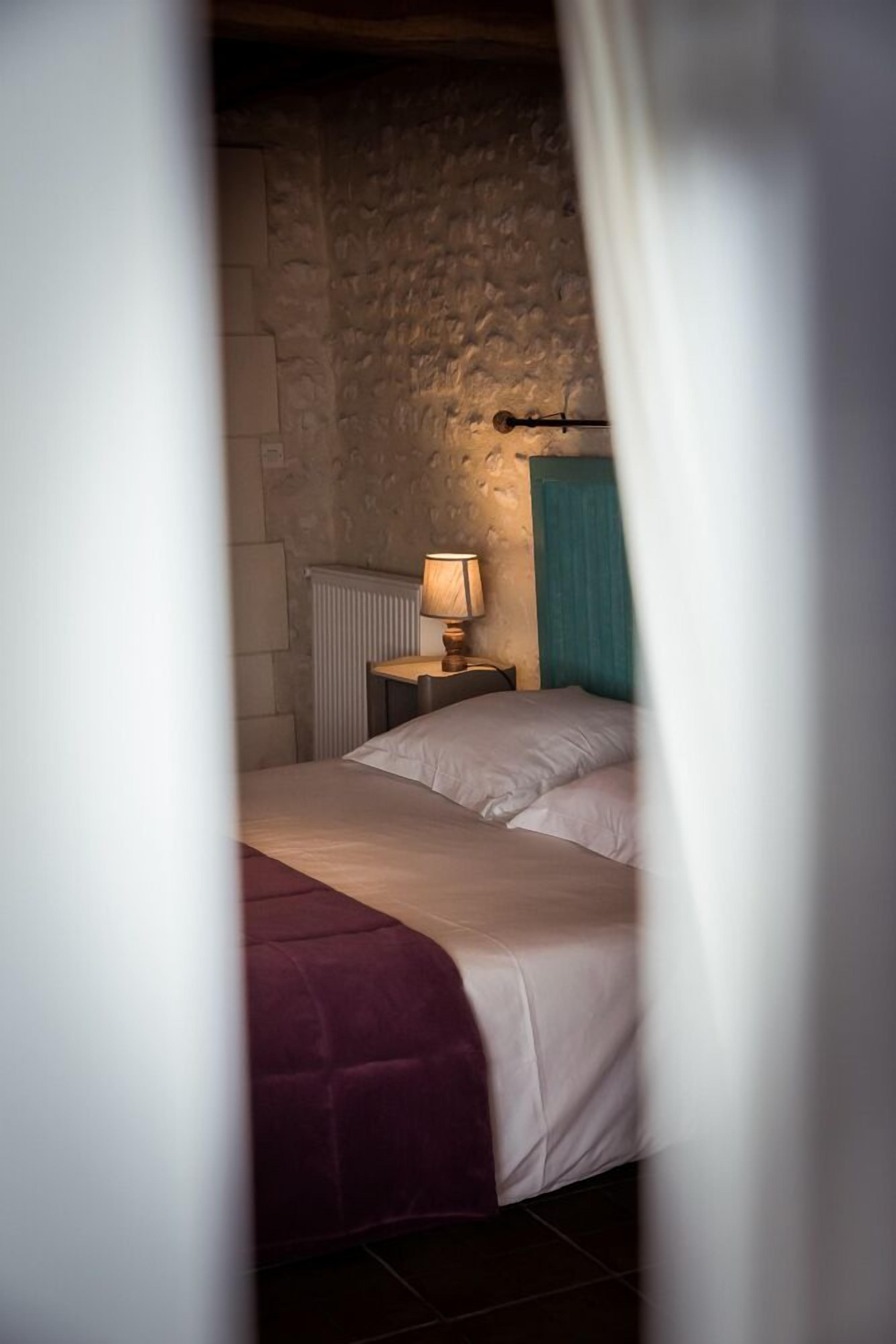 A bedroom with a stone wall, a bed with white bedding, a nightstand with a lamp, and a blue curtain.