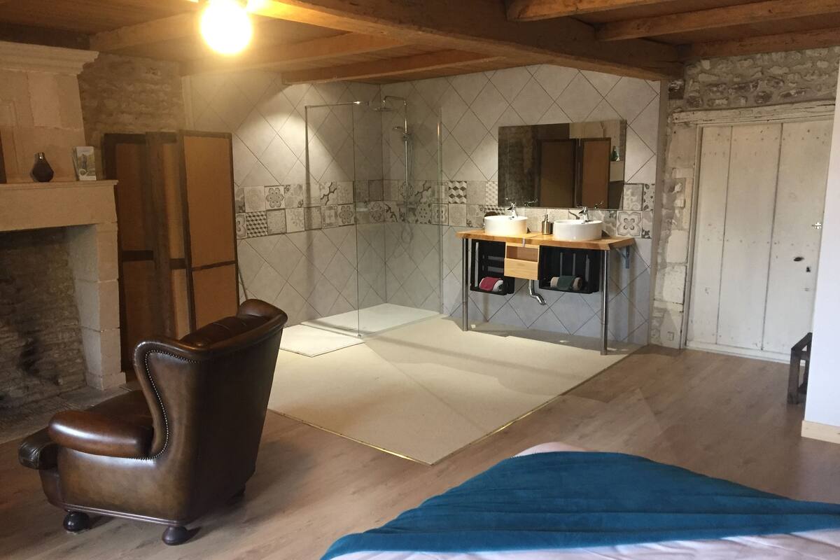 A room with a wooden ceiling, a fireplace, a leather chair, a bed with a blue blanket, and a bathroom with a shower and two sinks.