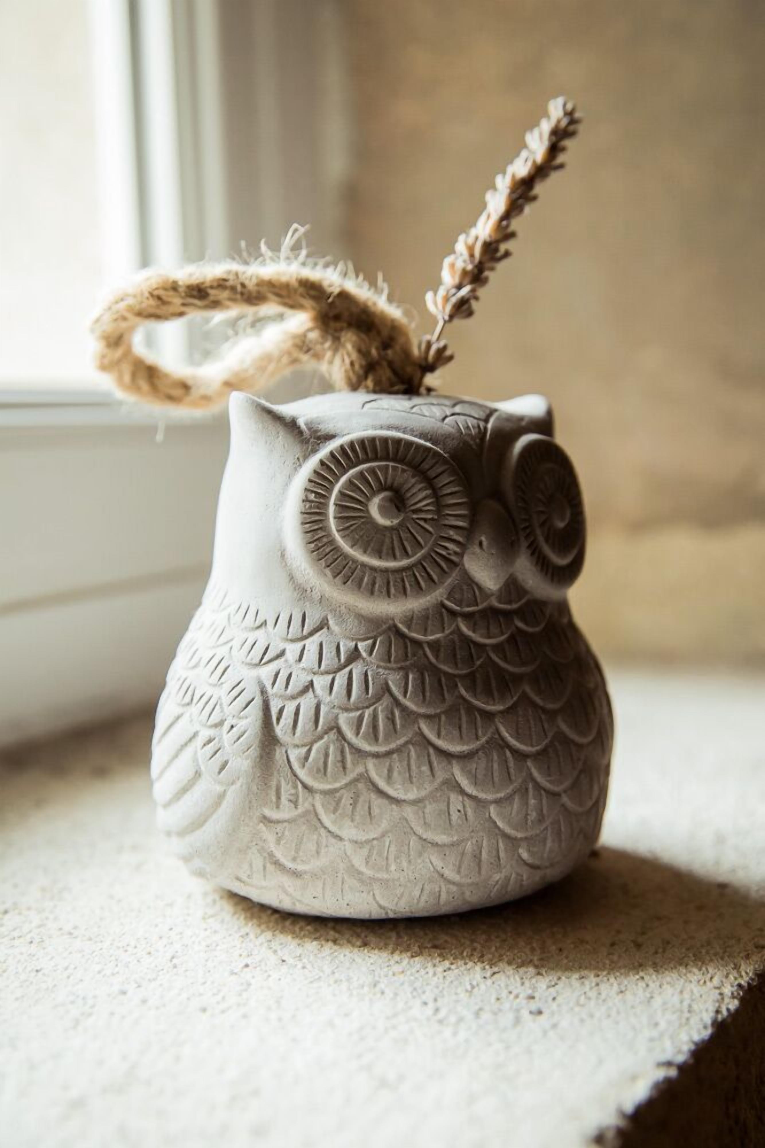 A ceramic owl-shaped container with a rope handle, placed on a textured surface near a window.