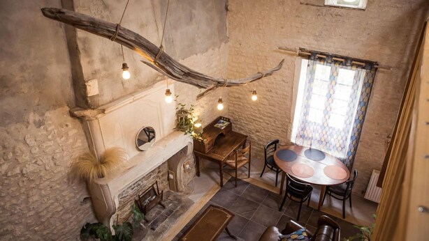 Un interior rústico con paredes de piedra vista, una mesa de comedor de madera con sillas, una chimenea y una lámpara colgante hecha con una rama.