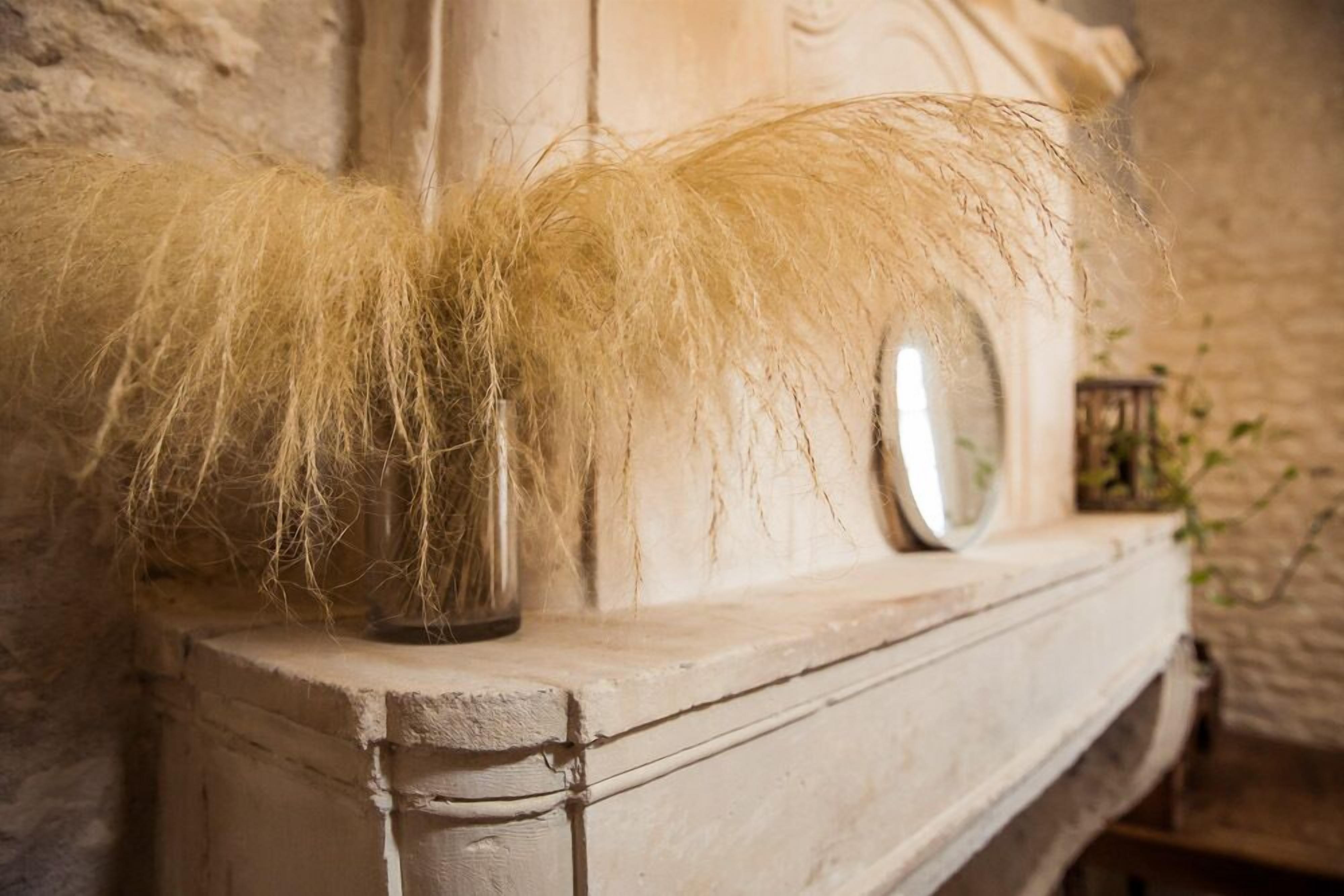 A rustic stone wall with a wooden shelf holding a vase of dried grass and a round mirror.