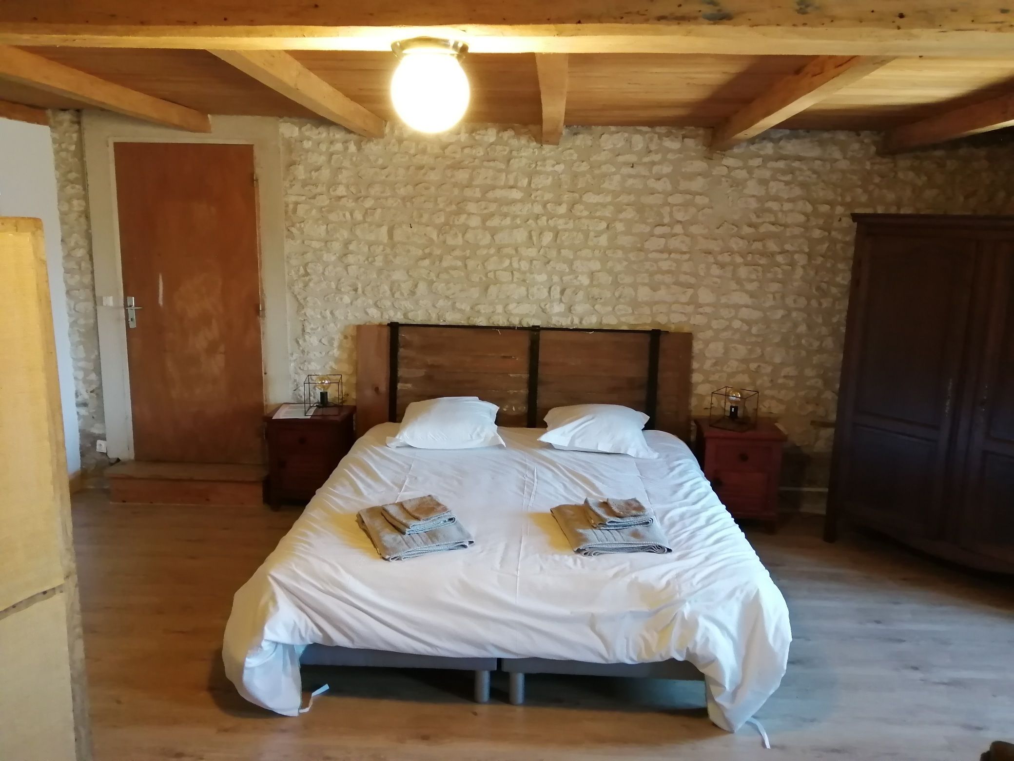 A bedroom with a stone wall, wooden ceiling beams, a bed with white bedding and towels, two bedside tables with lamps, and a wardrobe.