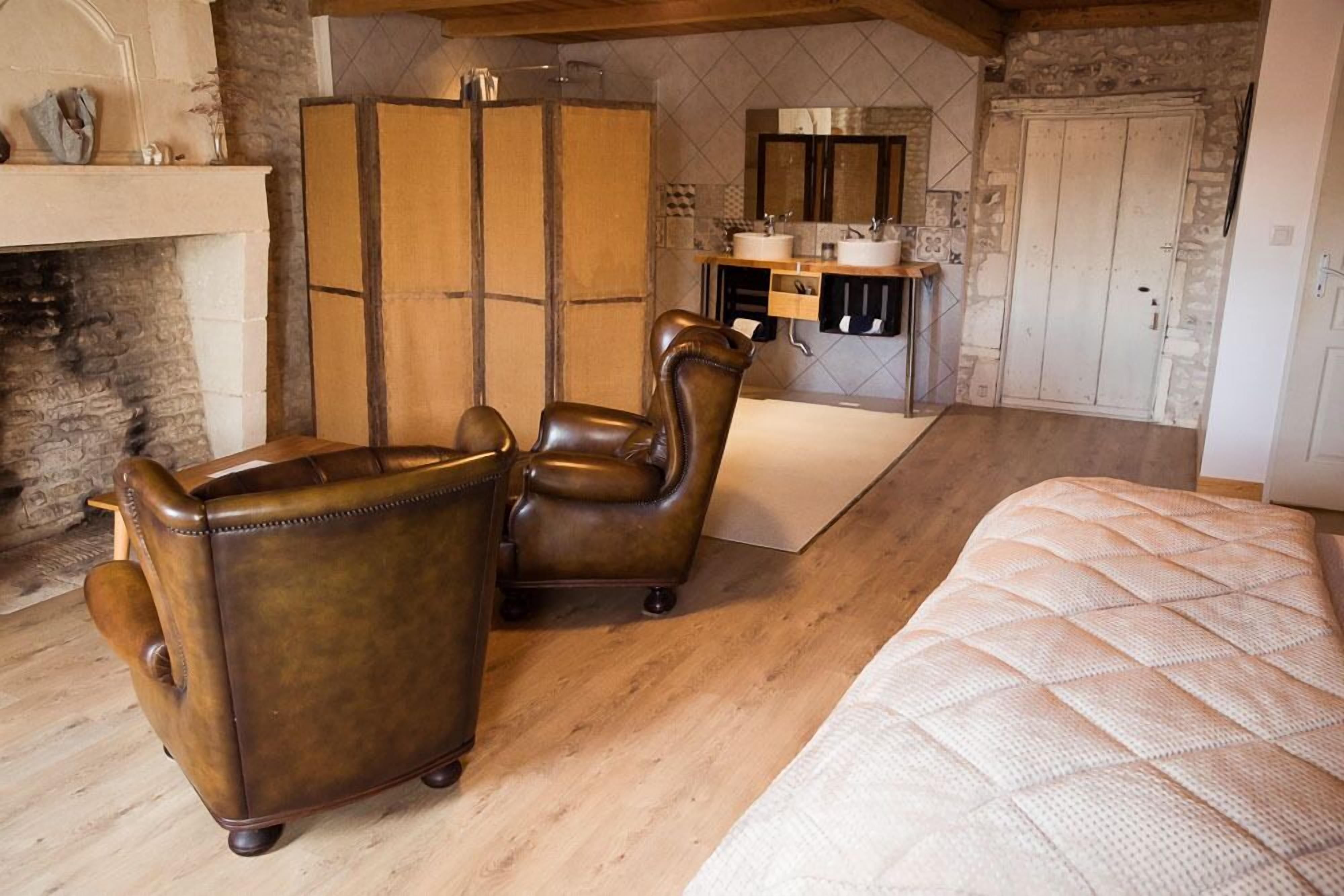 A bedroom with a large bed, two leather armchairs, a wooden floor, a stone wall, a fireplace, and a bathroom with a mirror and sink.