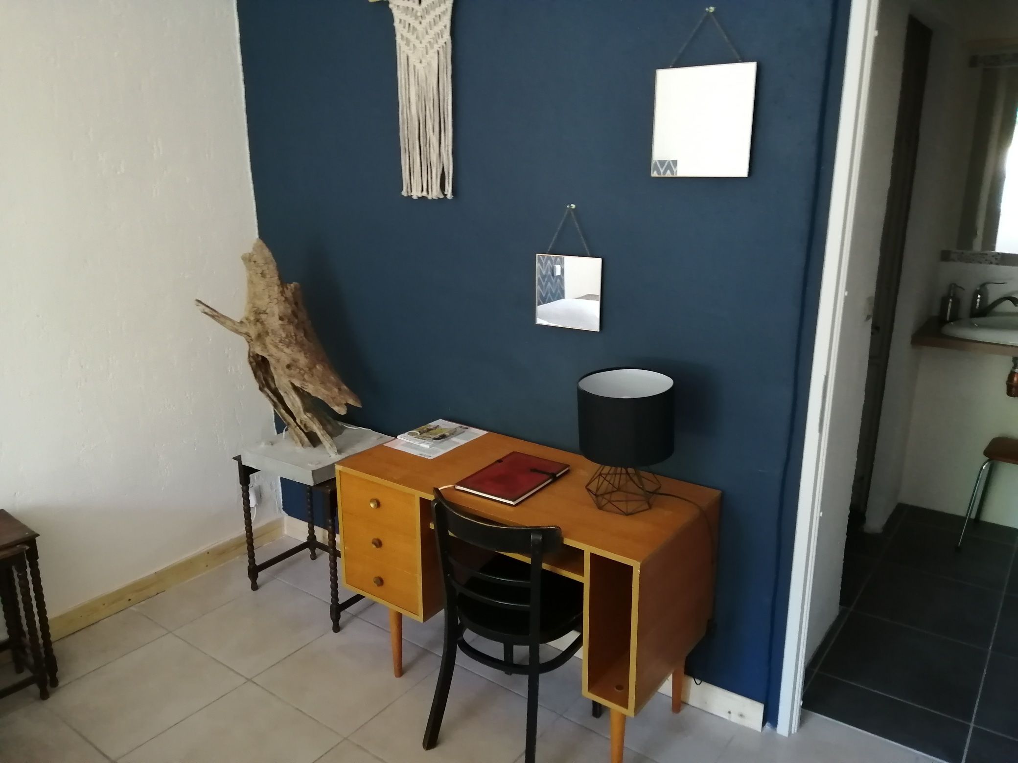 A small, compact room with a desk, chair, and a lamp. There is a mirror on the wall and a piece of artwork above the desk.