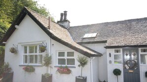 A white cottage with a steeply pitched roof, a front door with a heart-shaped decoration, and flower boxes on the windowsills.