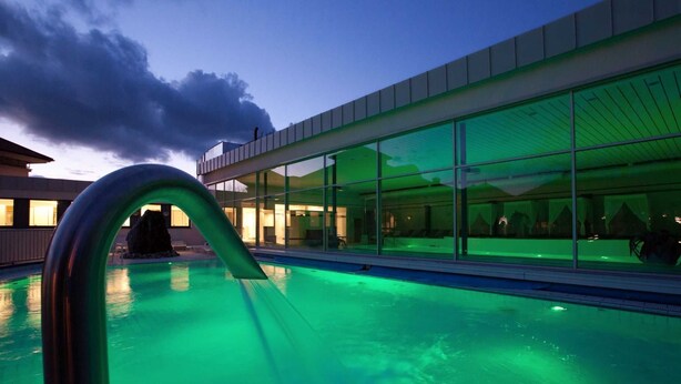 Un edificio moderno con grandes ventanales que reflejan el cielo y un área de piscina iluminada en verde.