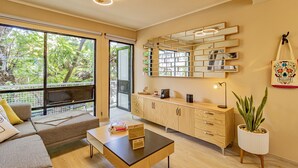 Una sala de estar moderna con un sofá, una mesa de centro y un espejo grande. Hay una planta sobre un pedestal y una ventana con vista a árboles.