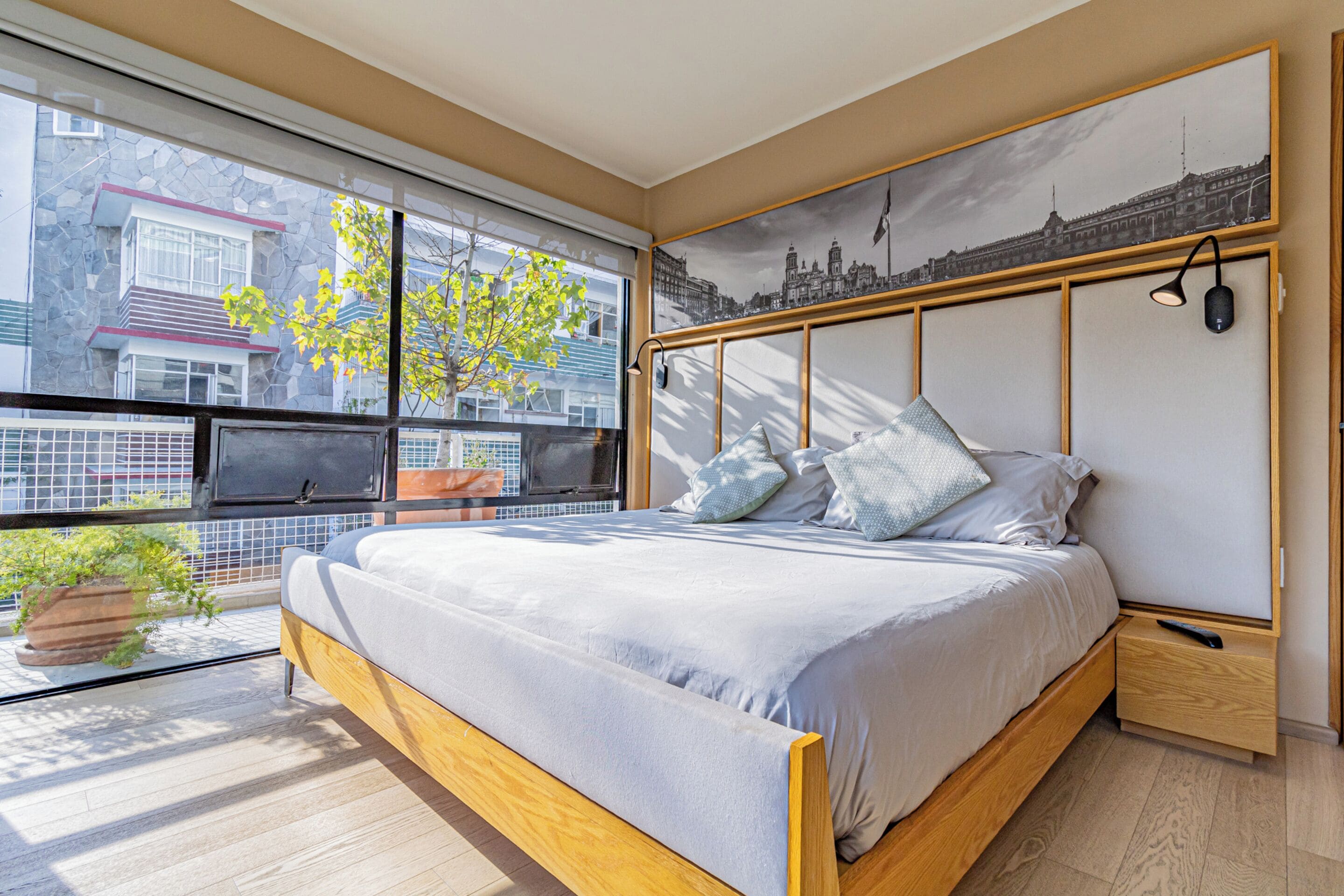 A modern bedroom with a large bed, a cityscape mural, a potted plant, and a window with a view of buildings.