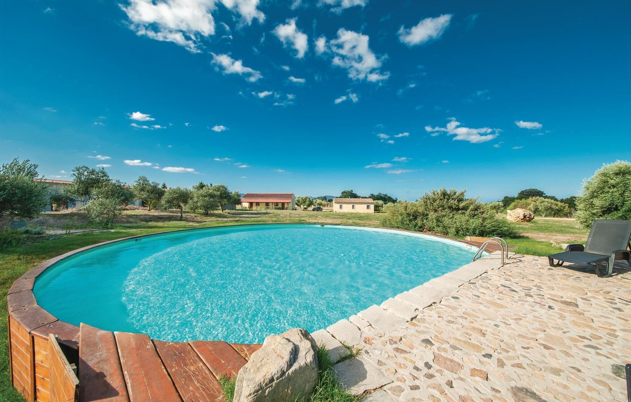 A swimming pool with a wooden deck and stone patio, surrounded by trees and a clear blue sky.