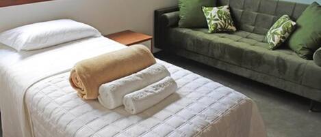 A neatly made bed with a white comforter and two rolled towels, a wooden nightstand, and a green sofa with patterned cushions.