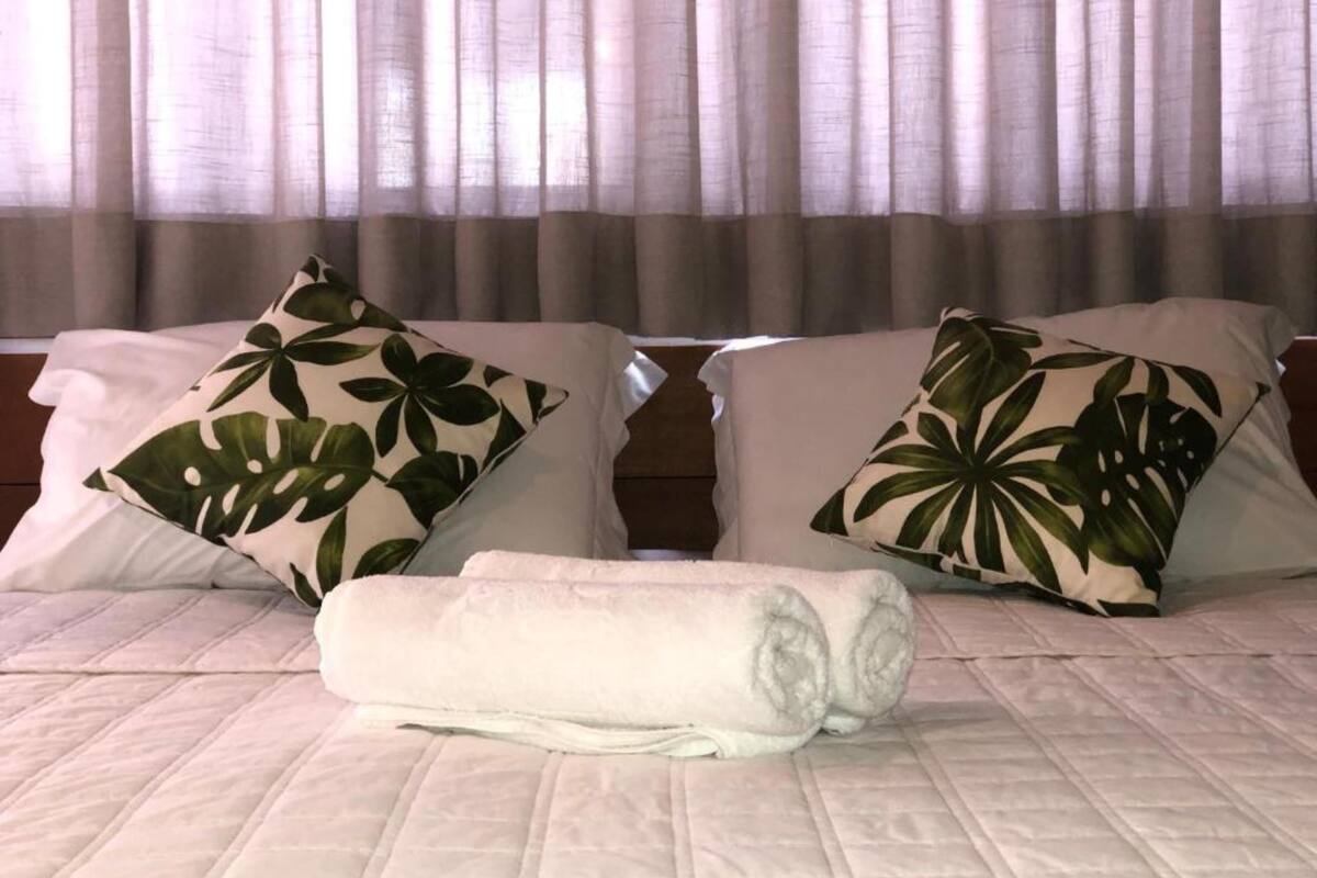 A bed with a quilted cover, two pillows with a tropical leaf pattern, and a rolled white towel.